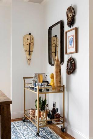 бели кухињски ормари, беле плоче, дрвени трпезаријски сто, дрвене барске столице, зидна уметност, колица са златним полугама